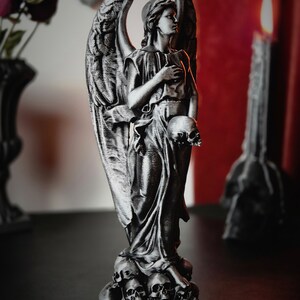 May include: A silver-colored angel statue with detailed wings and flowing robes, holding a skull. The statue stands atop a base of skulls, with a solemn expression. Gothic home decor.