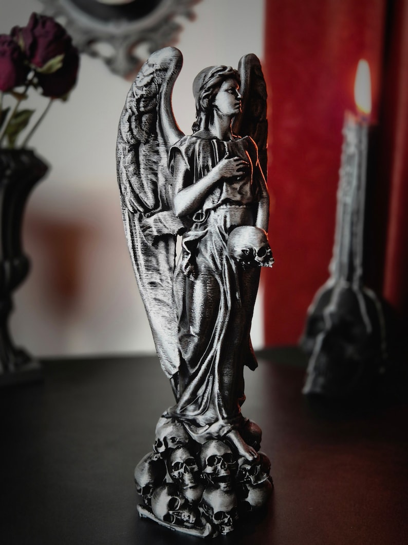 May include: A silver-colored angel statue with detailed wings and flowing robes, holding a skull. The statue stands atop a base of skulls, with a solemn expression. Gothic home decor.