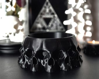 May include: A black bowl shaped like a ring of skulls. The bowl is made of a smooth, shiny material and has a slightly raised lip.