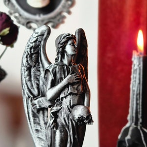 May include: A detailed, silver-colored angel statue holding a skull, with large wings and draped clothing. A lit black candle and a vase with dried flowers are in the background. The scene has a gothic theme.