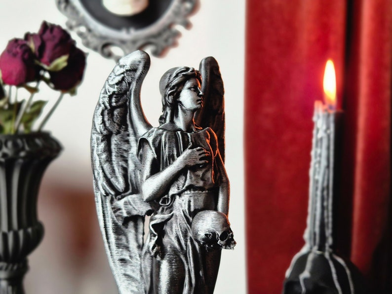 May include: A detailed, silver-colored angel statue holding a skull, with large wings and draped clothing. A lit black candle and a vase with dried flowers are in the background. The scene has a gothic theme.