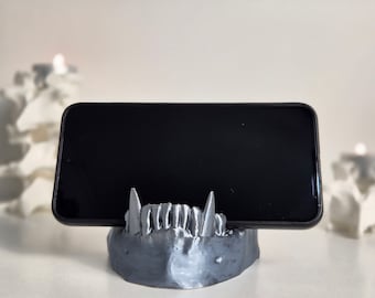 May include: A gray 3D printed phone stand shaped like a set of teeth. The stand is holding a black smartphone.