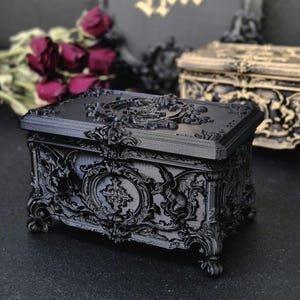 May include: A rectangular black box with intricate relief carvings featuring ornate scrollwork, a central cross emblem, and winged figures. The lid displays a circular decorative motif, and the base is supported by four small, detailed feet.