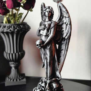 May include: A silver-colored angel statue holding a skull, standing atop a pile of skulls. The angel has large wings and is dressed in a flowing gown. A black vase with dried red roses is in the background.