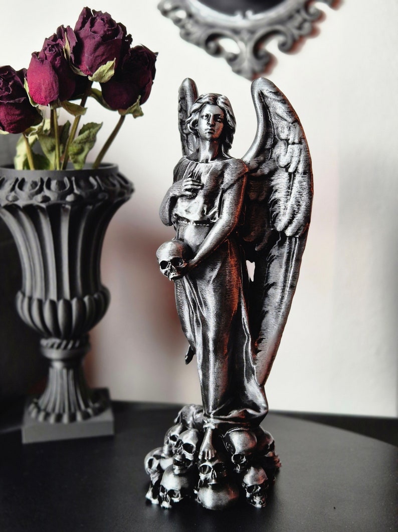 May include: A silver-colored angel statue holding a skull, standing atop a pile of skulls. The angel has large wings and is dressed in a flowing gown. A black vase with dried red roses is in the background.
