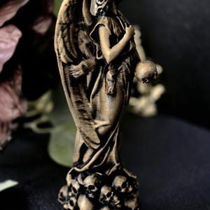 May include: A bronze-colored angel statue with detailed wings, holding a skull. The base of the statue is adorned with multiple skulls. The angel is wearing a draped garment.