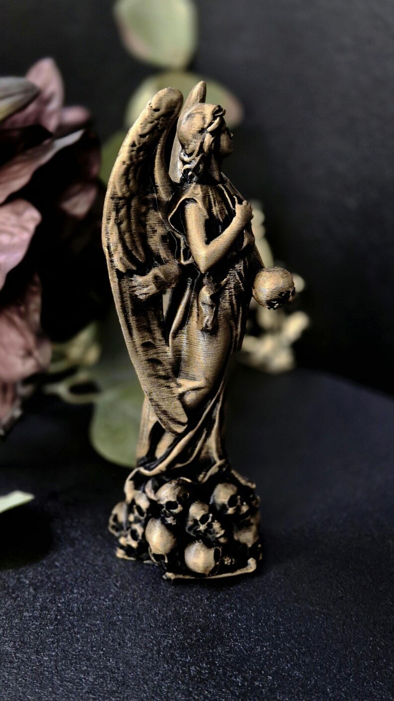 May include: A bronze-colored angel statue with detailed wings, holding a skull. The base of the statue is adorned with multiple skulls. The angel is wearing a draped garment.