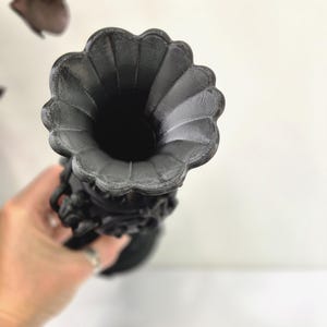 May include: A matte black vase featuring a wide, scalloped, flower-shaped rim and a textured, ornate body with intricate relief detailing. The vessel has a deep, hollow interior and a cylindrical neck that tapers towards the base.