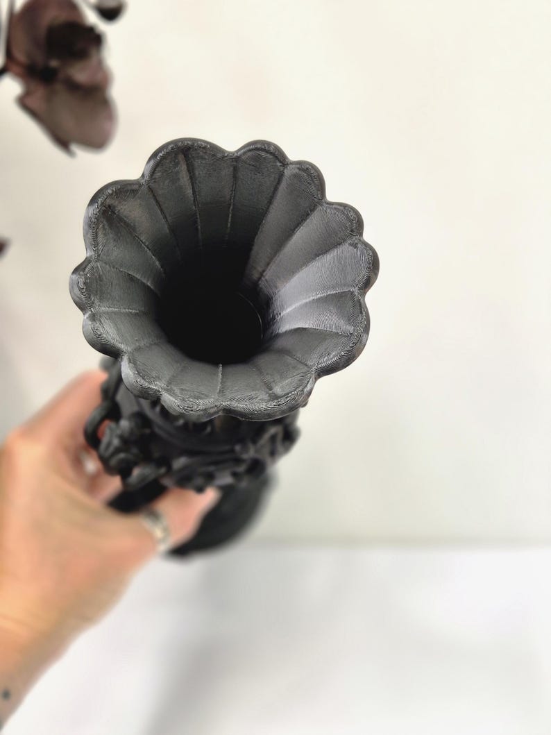 May include: A matte black vase featuring a wide, scalloped, flower-shaped rim and a textured, ornate body with intricate relief detailing. The vessel has a deep, hollow interior and a cylindrical neck that tapers towards the base.