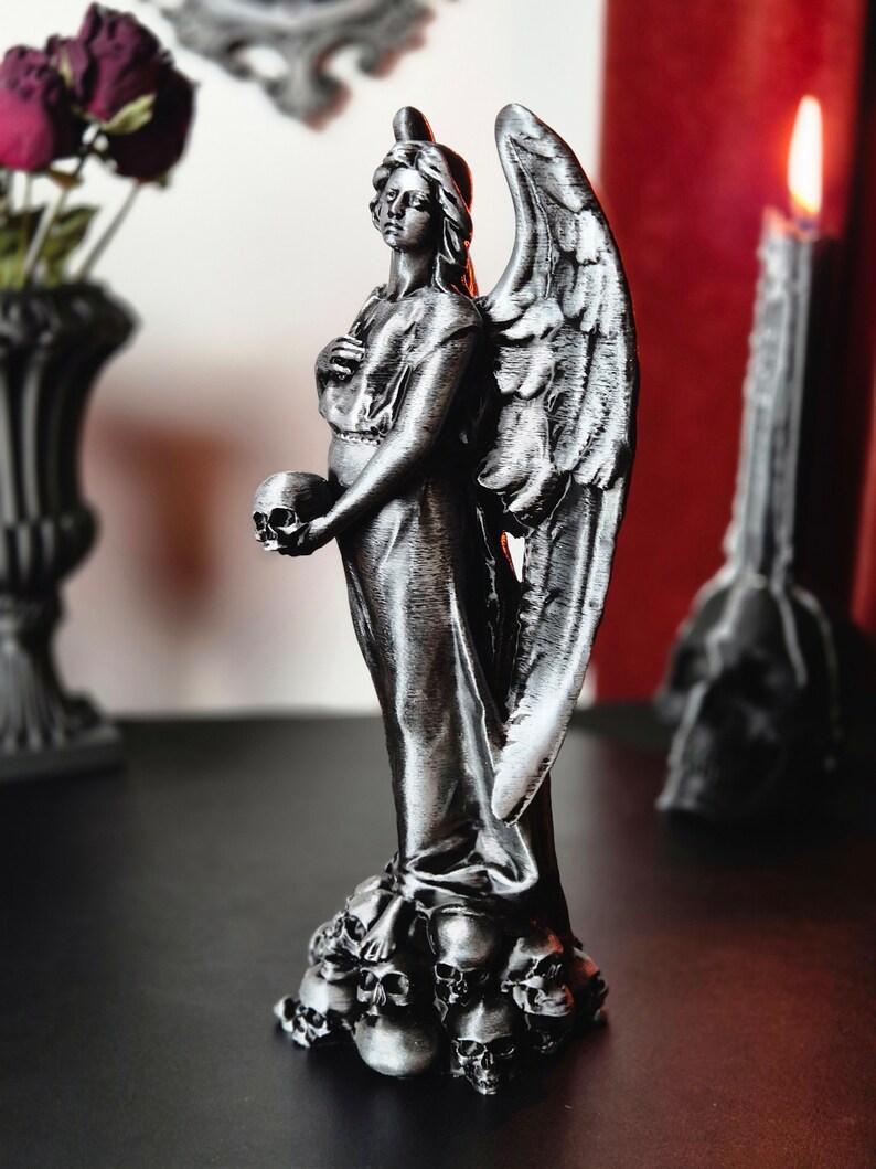 May include: A silver-colored angel statue holding a skull, with detailed wings and a flowing gown. The statue stands atop a base of small skulls. A candle and a vase of dark roses are in the background.
