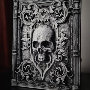 May include: A black and white 3D printed skull with ornate details, set against a background of a decorative book cover with gothic script. The book cover is made of a textured material and has a raised design.