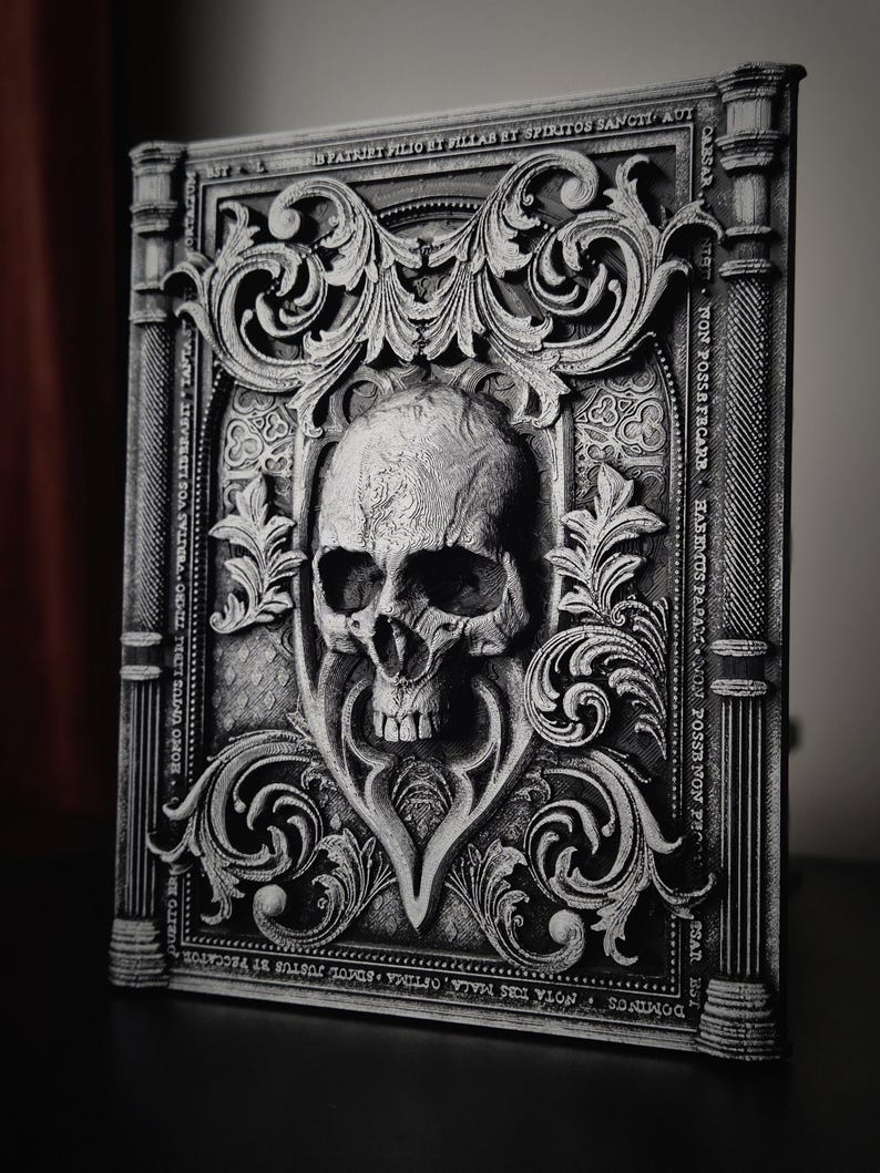 May include: A black and white 3D printed skull with ornate details, set against a background of a decorative book cover with gothic script. The book cover is made of a textured material and has a raised design.