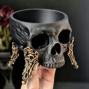 May include: A matte black skull-shaped planter or bowl featuring a hollowed-out top and three ornate, gold-colored decorative legs with intricate scrollwork detailing attached to the base of the skull, providing a stable foundation for the object.