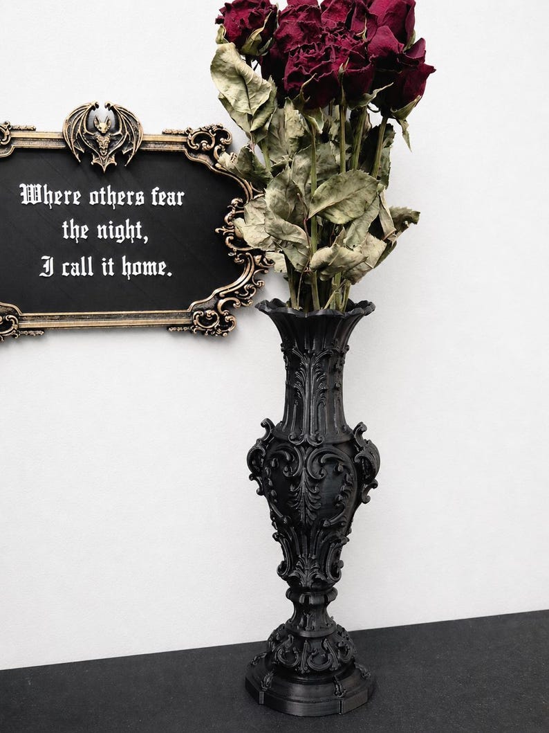 May include: A tall black vase with ornate scrollwork and a flared rim, holding a bouquet of deep red dried roses. Beside it hangs a rectangular black sign with a gold-toned frame, a bat emblem, and the text: Where others fear the night, I call it home.
