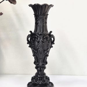 May include: A matte black vase featuring an ornate, raised relief pattern with intricate scrollwork and acanthus leaf motifs. The vessel has a flared, scalloped rim, a slender neck, a bulbous body, and a wide, circular base with detailed carvings.