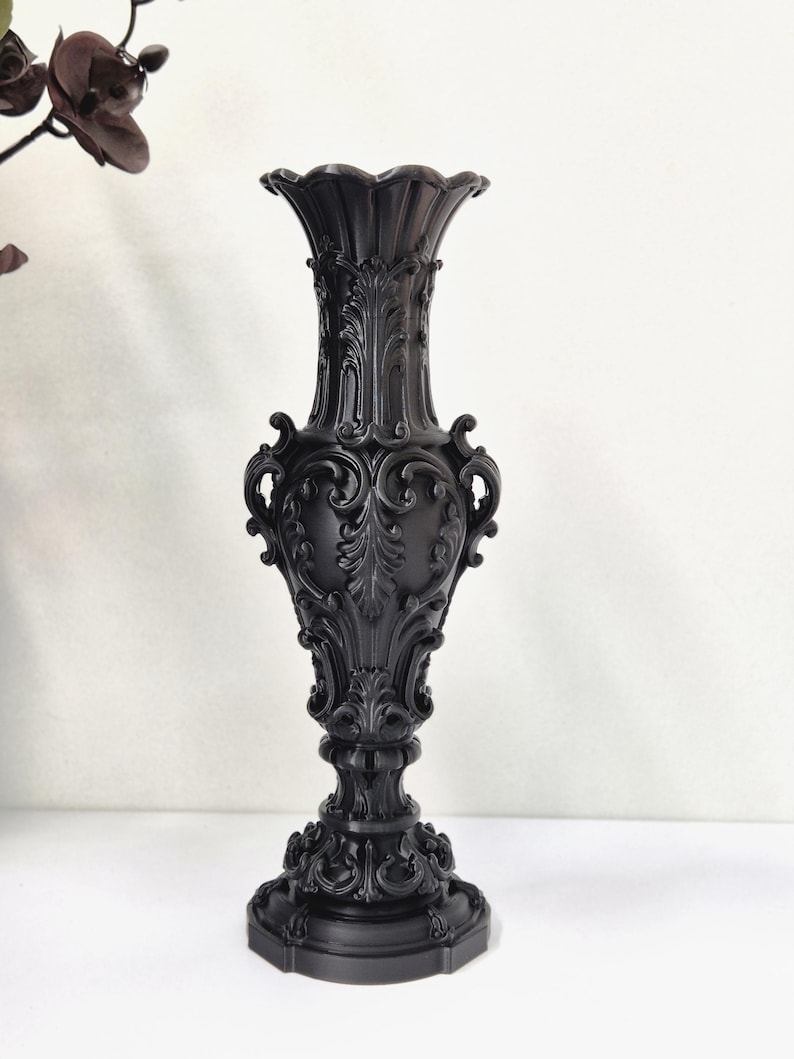 May include: A matte black vase featuring an ornate, raised relief pattern with intricate scrollwork and acanthus leaf motifs. The vessel has a flared, scalloped rim, a slender neck, a bulbous body, and a wide, circular base with detailed carvings.