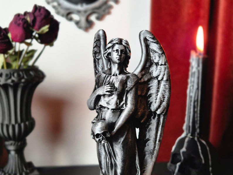 May include: A silver-colored angel statue with large wings, holding a skull. The statue is placed near a lit candle and a vase with dried flowers. The scene has a gothic or dark aesthetic.
