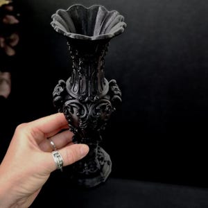 May include: A matte black vase featuring an ornate, raised relief pattern across its surface. The vessel has a wide, scalloped rim, a slender neck, and a bulbous body with decorative scrollwork details, resting on a sturdy, circular base.