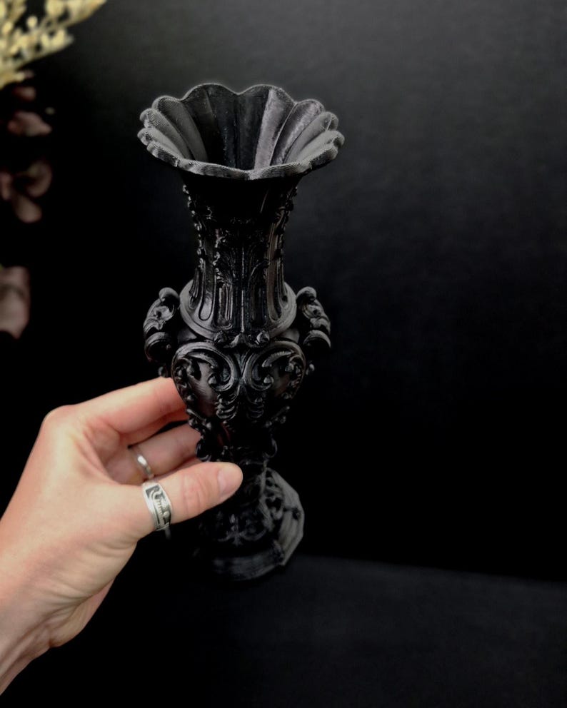 May include: A matte black vase featuring an ornate, raised relief pattern across its surface. The vessel has a wide, scalloped rim, a slender neck, and a bulbous body with decorative scrollwork details, resting on a sturdy, circular base.