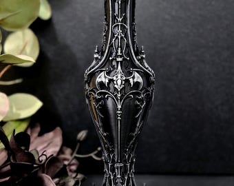 May include: A tall, slender black vase featuring intricate silver-toned relief detailing. The design includes symmetrical patterns, pointed architectural elements, and a central bat-like motif. The vase has a flared rim and a hexagonal base.