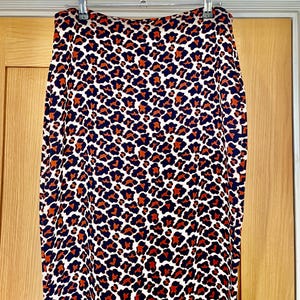 May include: A pencil skirt with a leopard print pattern. The skirt features a white background with dark navy blue and orange spots. The skirt is hanging on a silver hanger against a light brown wooden door.