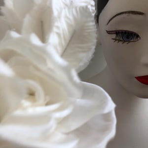 May include: A white fabric rose with multiple layers of petals. The rose is in focus and a white mannequin head with black hair and painted-on makeup is out of focus in the background.