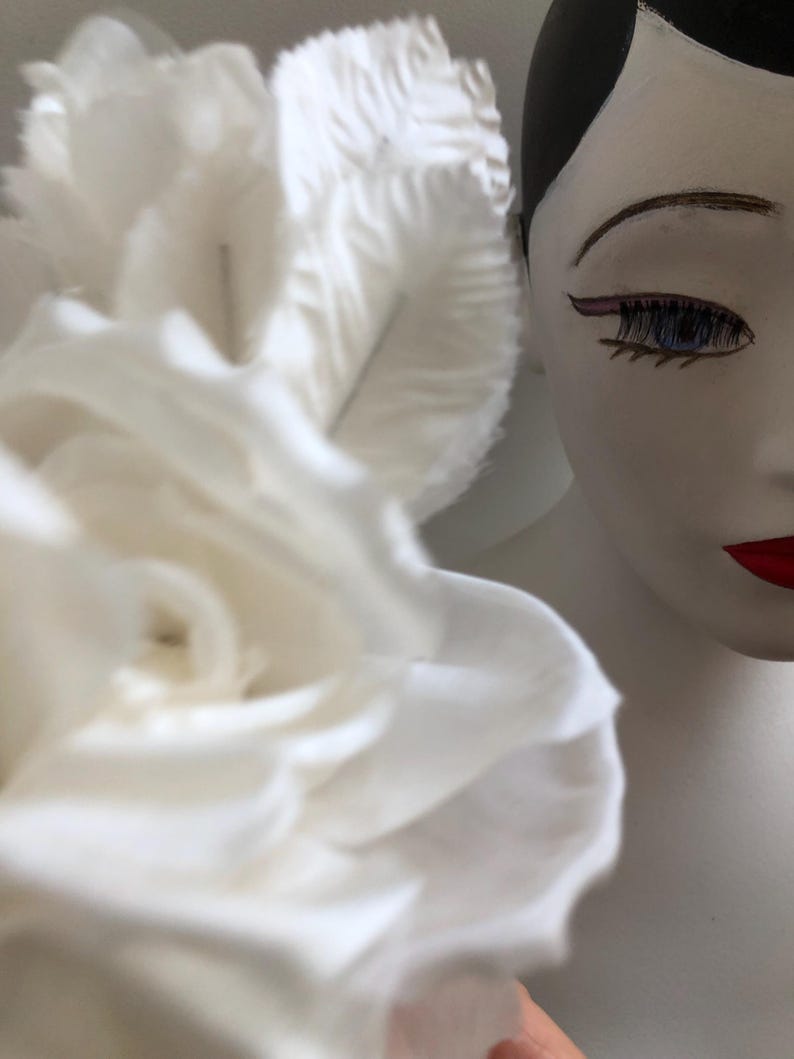 May include: A white fabric rose with multiple layers of petals. The rose is in focus and a white mannequin head with black hair and painted-on makeup is out of focus in the background.