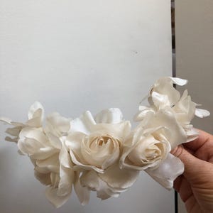 May include: A white flower crown made of silk roses. The crown is made of several roses, each with a different number of petals. The roses are arranged in a circle, with the petals overlapping.