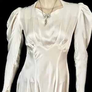 May include: Ivory-colored vintage dress with long sleeves and a V-neckline. The dress features a fitted bodice and a flowing skirt. A silver necklace with a pendant is draped on the dress. The dress has a classic, elegant design.