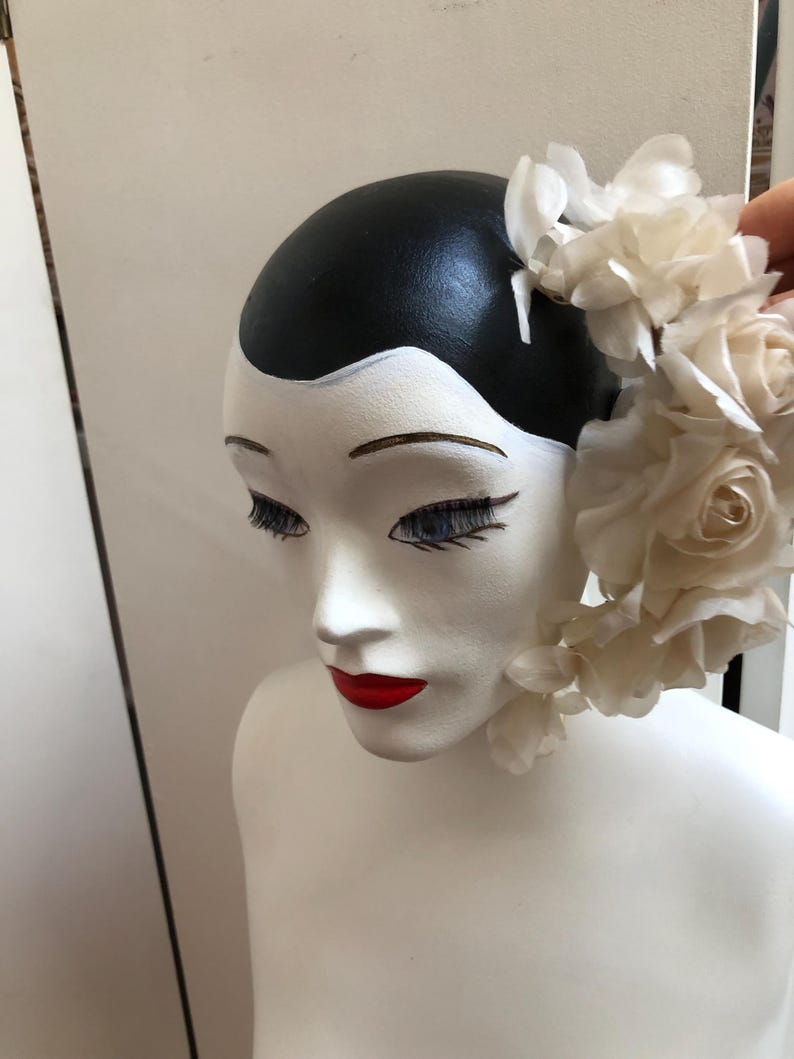 May include: A white mannequin head with black hair and red lips, wearing a white flower crown.