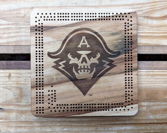 May include: A wooden coaster with a laser-engraved skull and crossbones design. The skull has a pirate hat with the letter "A" on it. The coaster has a border of small holes around the edge. The text "TM" is below the skull.