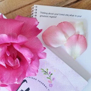 May include: A vibrant pink rose and scattered petals are next to a spiral-bound notebook. The notebook features the text "Thinking about your loved one, what is your greatest regret?" and a floral design. A journal is also visible.