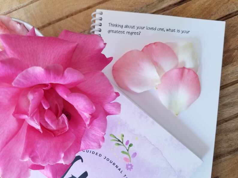 May include: A vibrant pink rose and scattered petals are next to a spiral-bound notebook. The notebook features the text "Thinking about your loved one, what is your greatest regret?" and a floral design. A journal is also visible.