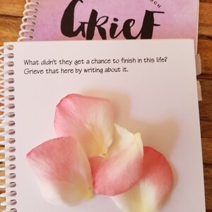 May include: A notebook with the word "Grief" on the cover. The open page has the text: "What didn't they get a chance to finish in this life? Grieve that here by writing about it." Pink and white rose petals are on the page.