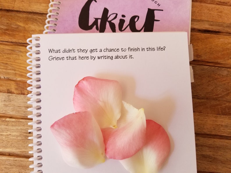 May include: A notebook with the word "Grief" on the cover. The open page has the text: "What didn't they get a chance to finish in this life? Grieve that here by writing about it." Pink and white rose petals are on the page.