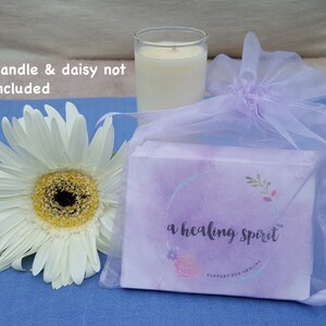 May include: A white daisy flower with a yellow center sits next to a clear glass candle holder with a lit candle. A small package wrapped in a sheer lavender bag is also present. The package has the text "a healing spirit".