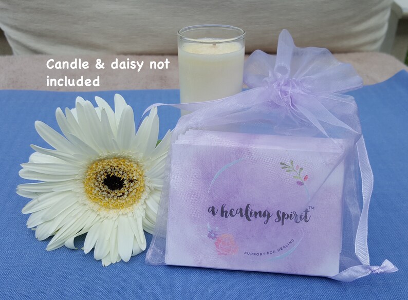 May include: A white daisy flower with a yellow center sits next to a clear glass candle holder with a lit candle. A small package wrapped in a sheer lavender bag is also present. The package has the text "a healing spirit".