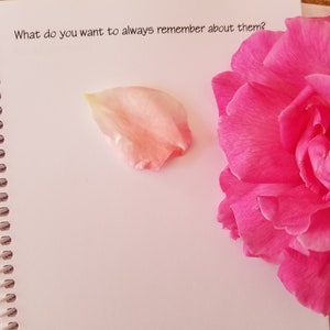 May include: An open spiral-bound notebook with the text "What do you want to always remember about them?" is shown. A pink flower and a single petal are on the page. The background is a wooden surface.