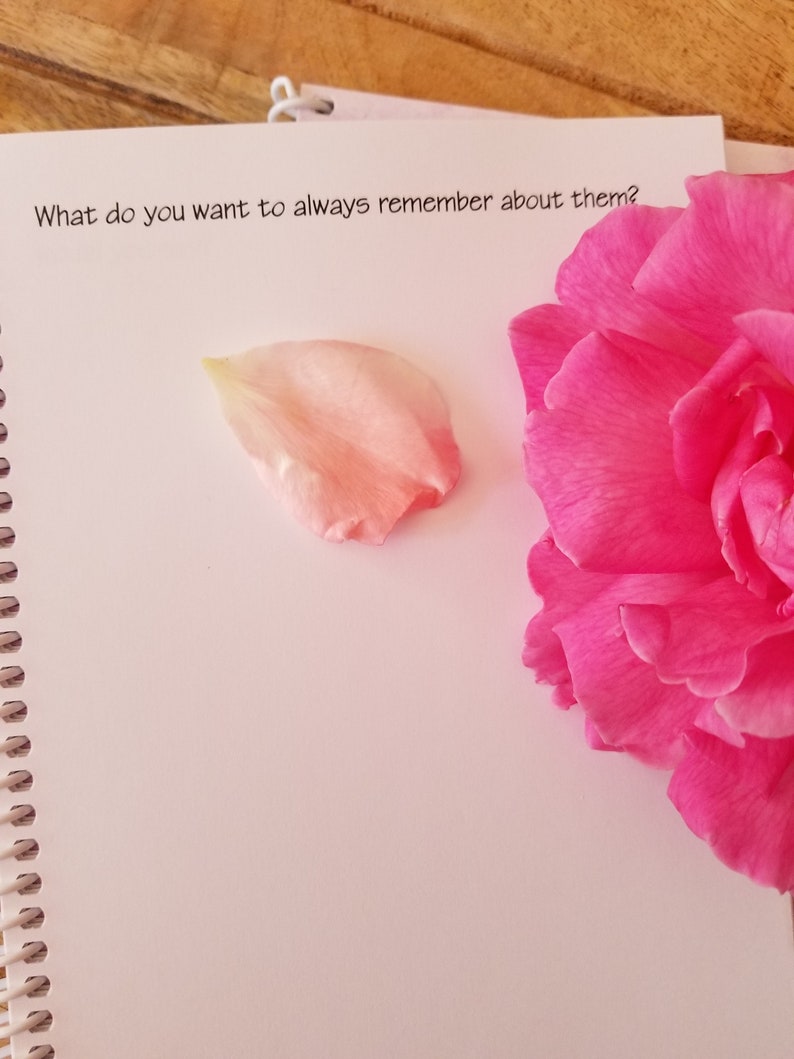 May include: An open spiral-bound notebook with the text "What do you want to always remember about them?" is shown. A pink flower and a single petal are on the page. The background is a wooden surface.