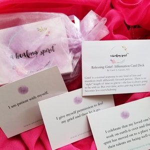 May include: A set of affirmation cards for releasing grief, with the text "a healing spirit" on the packaging. The cards have affirmations like "I am patient with myself" and "I give myself permission to feel my grief." The cards are on a pink background.