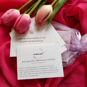 May include: A deck of affirmation cards for releasing grief, with text and a floral logo, is displayed with pink tulips and a sheer pink fabric. The cards are in a small, sheer, lavender bag. The text on the card reads: "I give myself permission to feel my grief and then let it go."