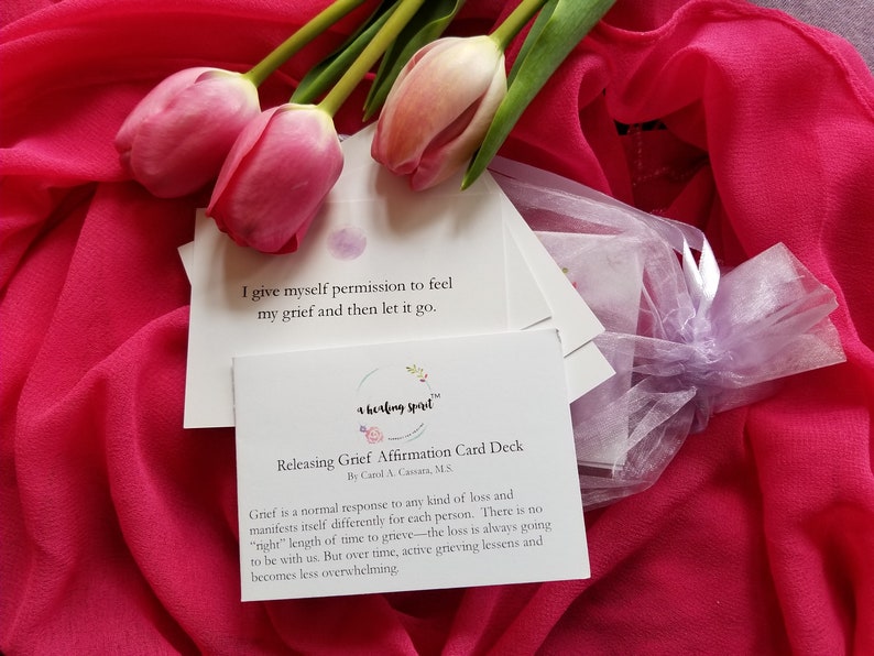 May include: A deck of affirmation cards for releasing grief, with text and a floral logo, is displayed with pink tulips and a sheer pink fabric. The cards are in a small, sheer, lavender bag. The text on the card reads: "I give myself permission to feel my grief and then let it go."