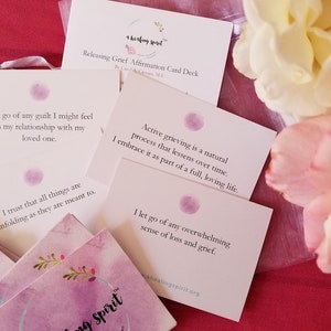 May include: A deck of affirmation cards titled "Releasing Grief Affirmation Card Deck" with text and a watercolor design. The cards are white with purple accents and floral details. The image includes white and pink flowers.