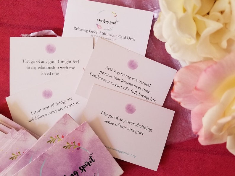 May include: A deck of affirmation cards titled "Releasing Grief Affirmation Card Deck" with text and a watercolor design. The cards are white with purple accents and floral details. The image includes white and pink flowers.