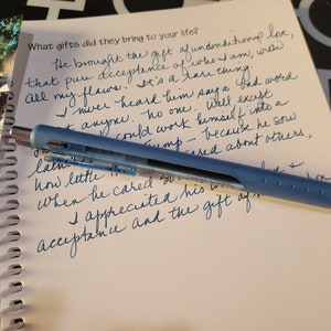 May include: A blue multi-pen lies on a spiral-bound notebook with handwritten text in blue ink. The text reads, "What gifts did they bring to your life?" A photograph of a person is in the background.