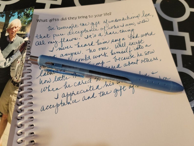 May include: A blue multi-pen lies on a spiral-bound notebook with handwritten text in blue ink. The text reads, "What gifts did they bring to your life?" A photograph of a person is in the background.