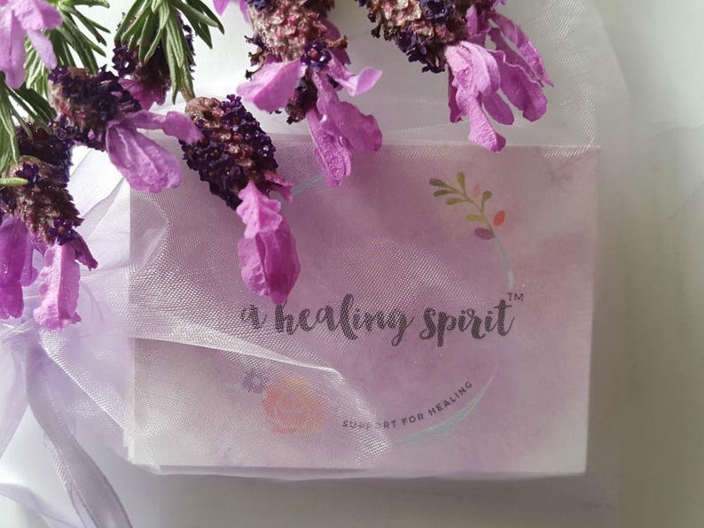 May include: A lavender-colored package wrapped in sheer white fabric, adorned with sprigs of purple lavender flowers. The package features the text "a healing spirit" and "support for healing" in a delicate script, suggesting a wellness product.