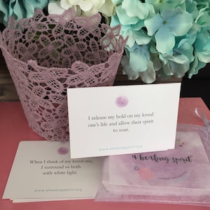 May include: A stack of white cards with text and a purple watercolor design, with a purple lace basket and a bouquet of flowers in the background. The text on the cards reads "I release my hold on my loved one's life and allow their spirit to soar."