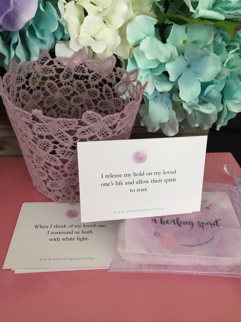 May include: A stack of white cards with text and a purple watercolor design, with a purple lace basket and a bouquet of flowers in the background. The text on the cards reads "I release my hold on my loved one's life and allow their spirit to soar."