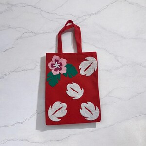May include: Red tote bag with a floral design. The bag features a pink and purple flower with green leaves, and white leaf-shaped accents. The bag has a handle and is made of a sturdy material.